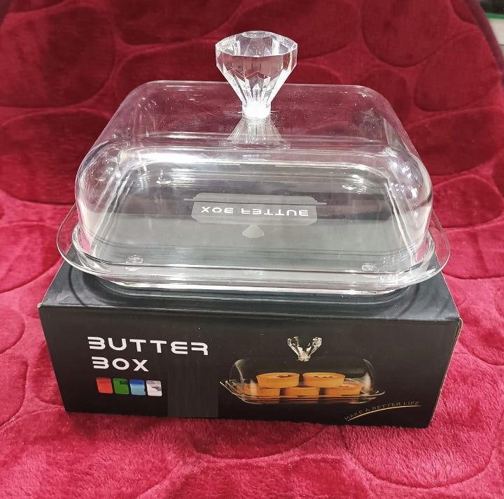 An acrylic butter dish with a plastic lid and knob, displayed on a red surface, with the product packaging visible in the background.