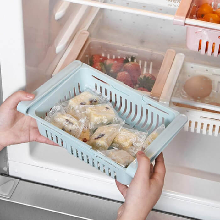 Person holding a blue plastic basket with wrapped food items in front of an open refrigerator.