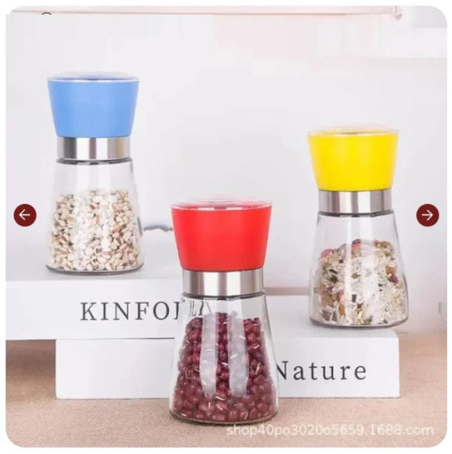 Elegant stainless steel pepper grinder with a clear glass body, shown set to a coarse grind, being used to season a dish of fresh food on a wooden counter.