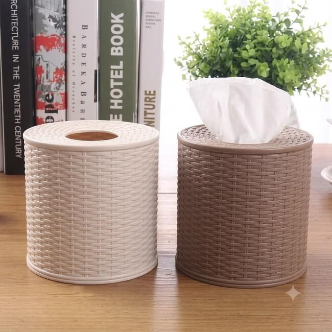 Two woven tissue holders on a wooden surface with books and a plant in the background.