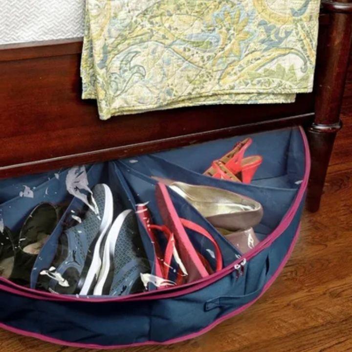 A navy blue shoe bag with pink trim and an internal organizer divider, shown on a wooden floor under a bed with various shoes stored inside.
