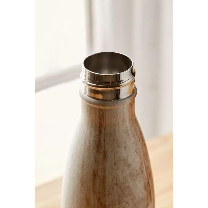 Close-up of a reusable bottle with a brushed metallic silver stainless steel rim and a textured beige-brown ceramic body, set against a soft-focus background of white curtains and a light wooden surface.