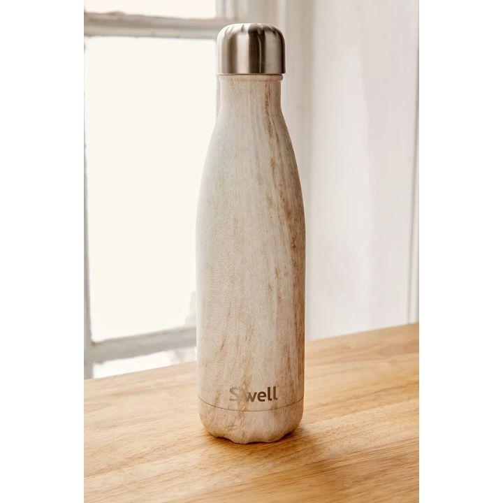 A reusable insulated water bottle with a light beige marble-look finish and a brushed silver metal cap, standing on a wooden tabletop in front of a softly blurred window.