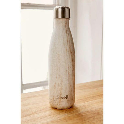 A reusable insulated water bottle with a light beige marble-look finish and a brushed silver metal cap, standing on a wooden tabletop in front of a softly blurred window.