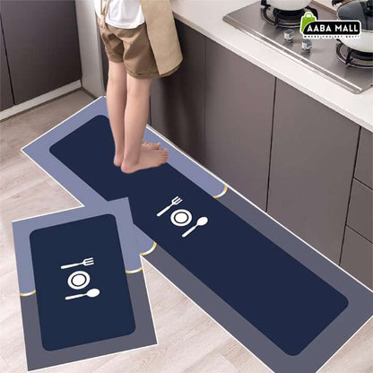 A rectangular kitchen mat with a dark navy blue center, gray border, and white table setting icon, shown in a kitchen with a child stepping on it. A smaller matching mat is displayed in the foreground, and an 'AABA MALL' logo appears in the top right corner.