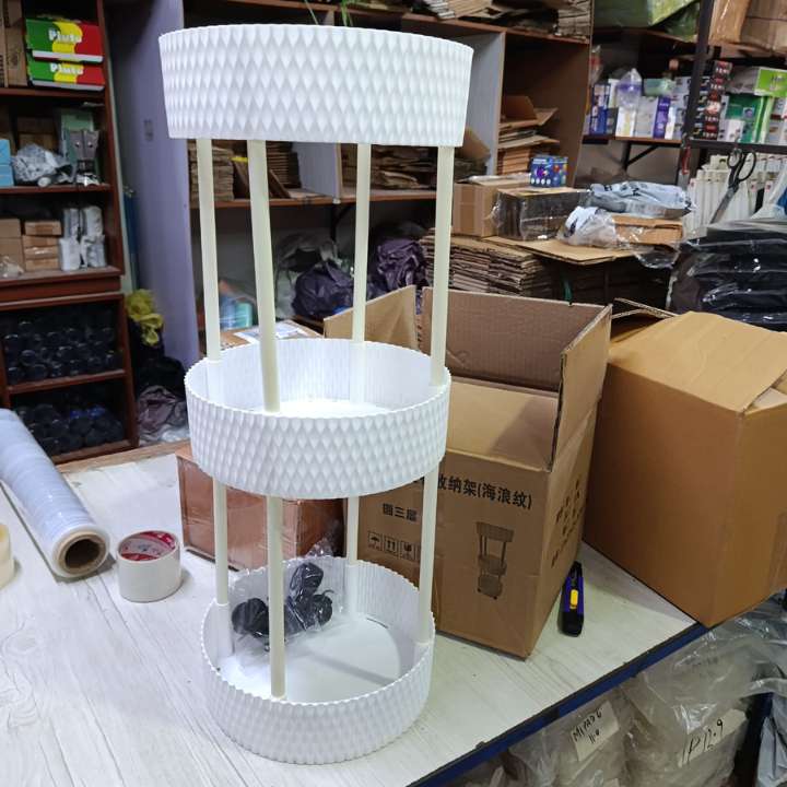A three-tier white plastic plant stand with a textured, diamond-patterned rim and vertical support rods, shown in a warehouse or storage room setting with cardboard boxes and shelves in the background.