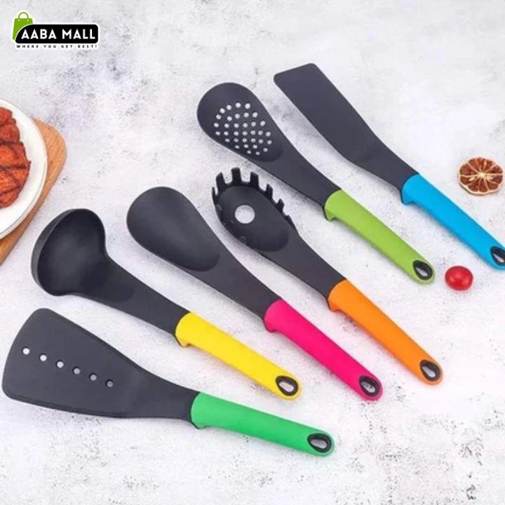 Six silicone kitchen utensils with black heads and brightly colored handles are arranged on a light textured surface. The utensils include a spatula, ladle, spaghetti server, whisk, and another flatturner.