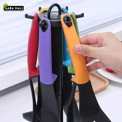A set of kitchen utensils with colorful handles in red, purple, blue, green, and orange, displayed on a black stand with a hand holding one of the orange handles. The AABA MALL logo is visible in the top-left corner.