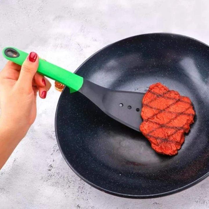 A hand with red nail polish holds a green-handled spatula lifting a grilled red steak from a black speckled frying pan on a light gray textured surface.