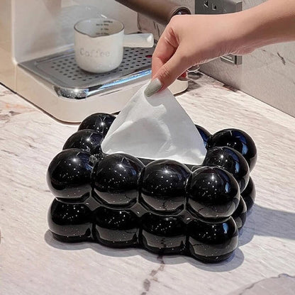 Black bubble-shaped tissue holder with a hand pulling out a tissue, placed on a marble surface next to a coffee machine.