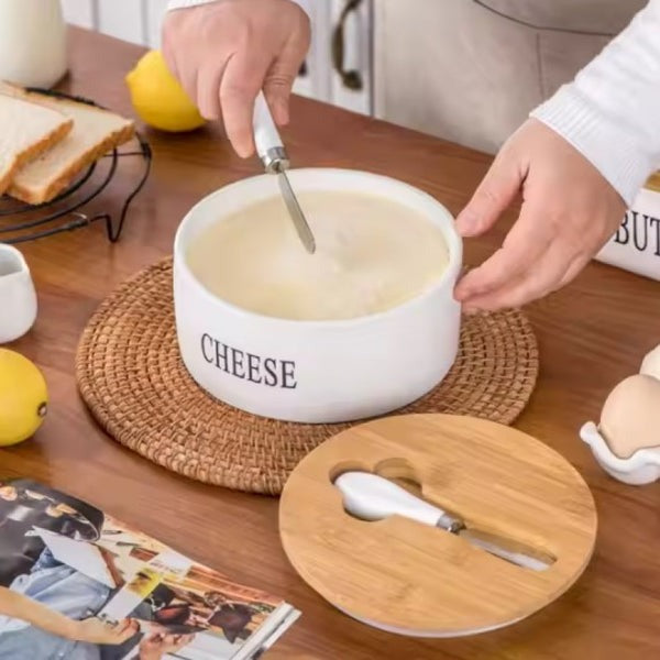 Elegant white ceramic cheese and butter storage box with a natural bamboo lid and three stainless steel serving knives displayed next to it on a wooden counter.