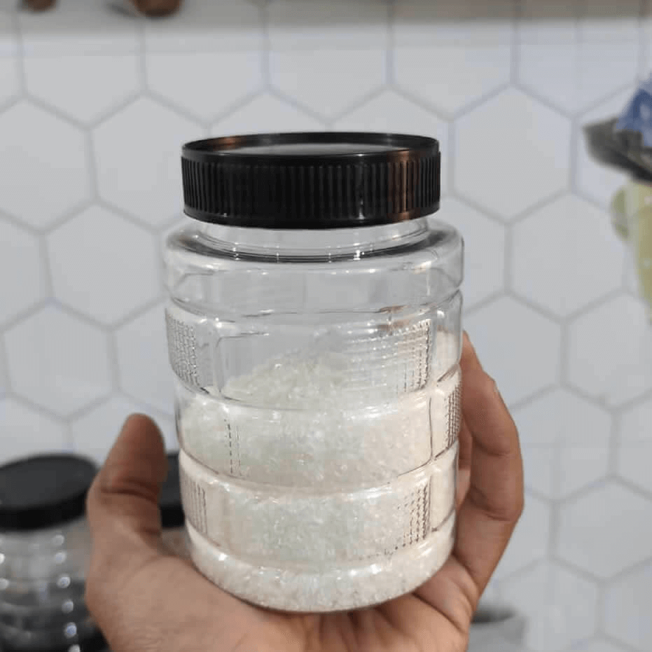Hand holding a clear jar with a black lid filled with sugar against a tiled wall background.