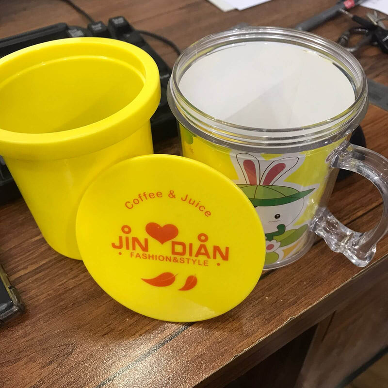 Yellow mug with a lid and clear mug with cartoon character on a wooden surface