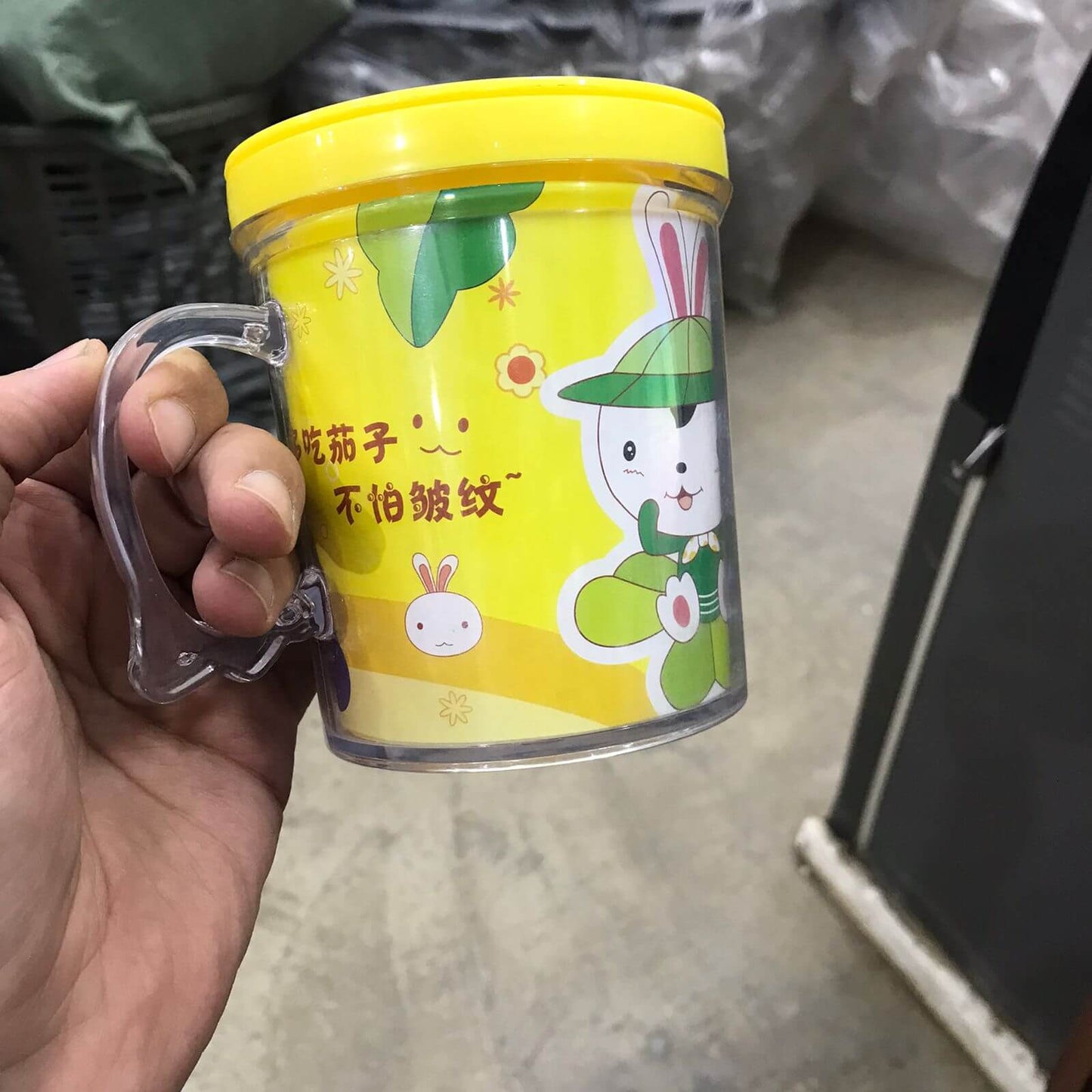 Hand holding a yellow mug with cartoon character design on a concrete floor.