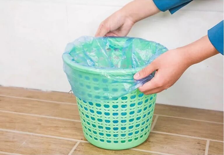 Person putting a plastic bag into a green laundry basket on a wooden floor.
