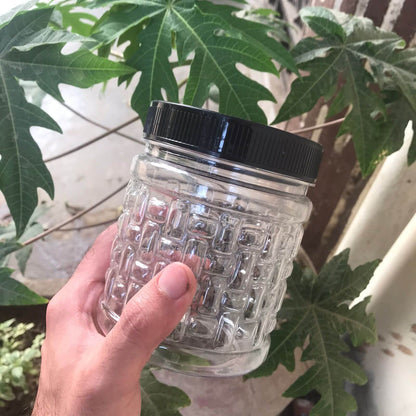 Clear textured hard plastic jar with black lid held by a hand against a leafy green background