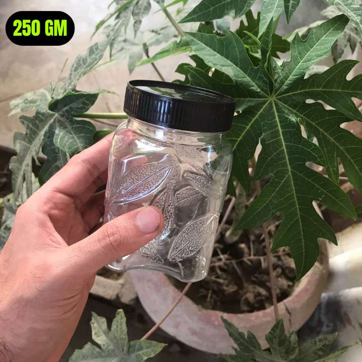Hand holding a clear jar with black lid containing dried leaves, with green leaves in the background.