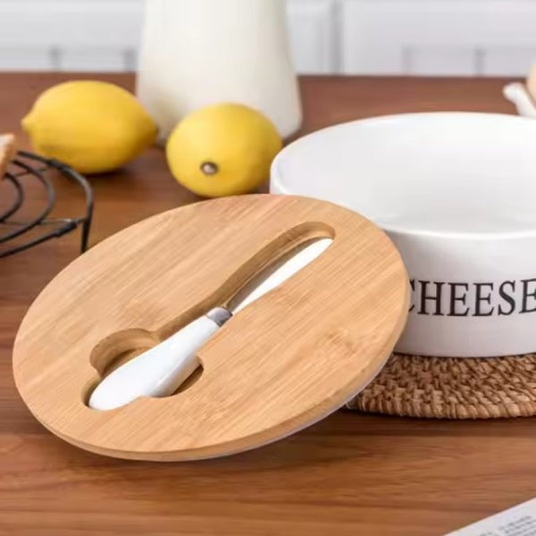Elegant white ceramic cheese and butter storage box with a natural bamboo lid and three stainless steel serving knives displayed next to it on a wooden counter.