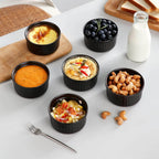 Assorted snacks in black ramekins on a white surface with a bottle of milk and bread in the background.