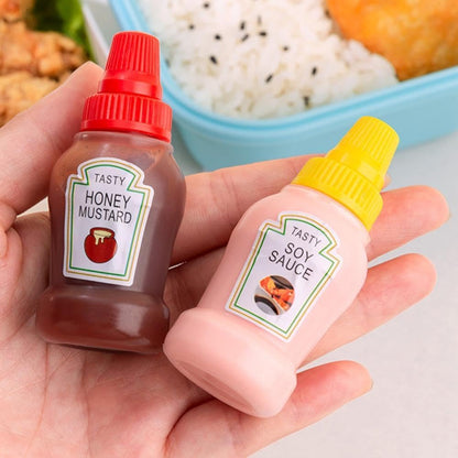 Set of two clear mini squeeze condiment bottles, one filled with red ketchup and one with white mayonnaise, next to a packed lunch box.