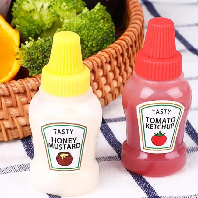 Set of two clear mini squeeze condiment bottles, one filled with red ketchup and one with white mayonnaise, next to a packed lunch box.