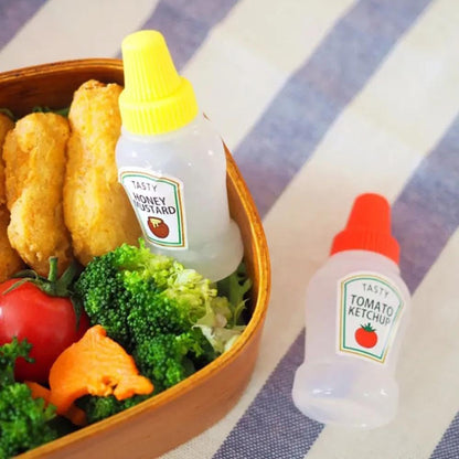 Set of two clear mini squeeze condiment bottles, one filled with red ketchup and one with white mayonnaise, next to a packed lunch box.