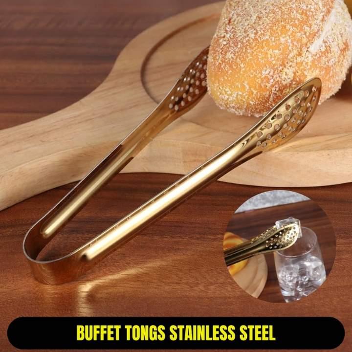 Gold buffet tongs on a wooden surface with bread and a glass of water.