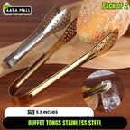 Gold and silver buffet tongs on a wooden surface with bread and a glass of water.