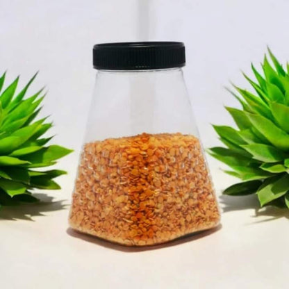 Clear container with black lid filled with orange seeds, placed between two green plants on a light background