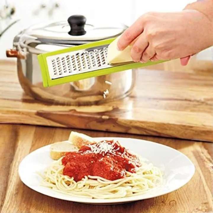 Person using a green box grater over a plate of spaghetti with a glass container in the background.