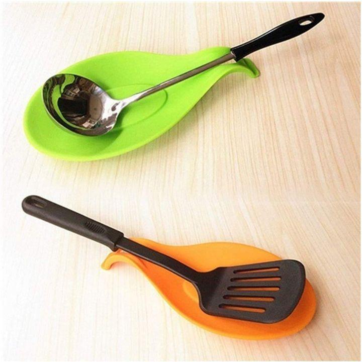Two silicone kitchen utensil holders on a light wood surface: a bright green holder with a stainless steel spoon, and an orange holder with a black spatula.