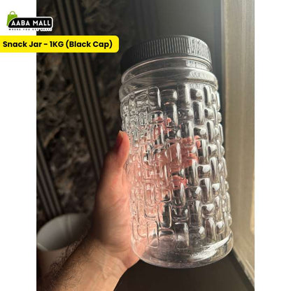 A hand holding a clear plastic snack jar with a textured embossed pattern and a black ribbed cap, set against a dark indoor background with a window light.
