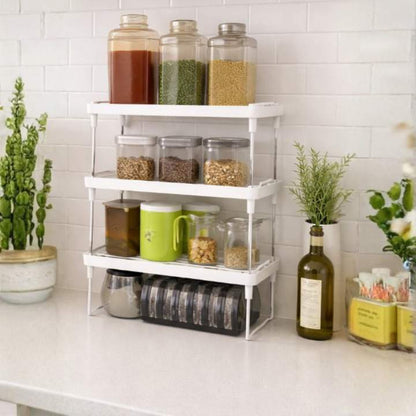 Kitchen counter with a white storage rack holding various jars and containers, with plants and bottles in the background.