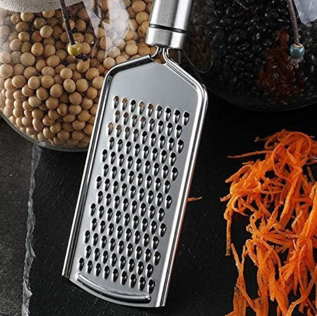 Close-up of a stainless steel handheld cheese grater/zester with razor-sharp micro-blades, shown zesting the bright yellow peel of a lemon on a cutting board.