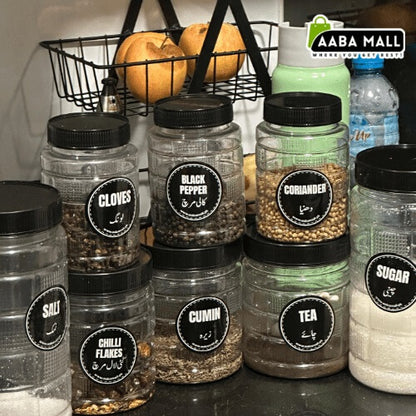 Clear cubic plastic storage jars with black lids arranged on a white surface with plants in the background