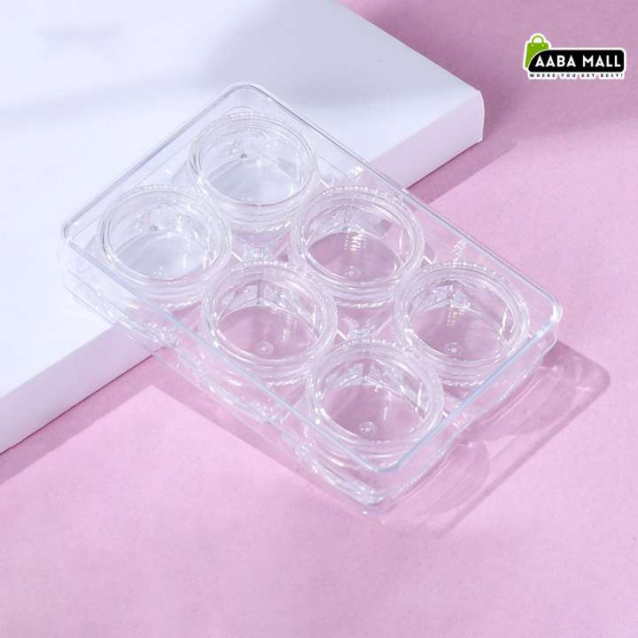 A clear plastic six-compartment tray, likely for contact lens packaging, resting on a white rectangular block against a soft pink background. An 'AABA MALL' logo is visible in the top right corner.