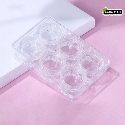 A clear plastic six-compartment tray, likely for contact lens packaging, resting on a white rectangular block against a soft pink background. An 'AABA MALL' logo is visible in the top right corner.