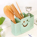 A green plastic cutlery holder with a smiley face design, containing various wooden utensils such as spoons and chopsticks.