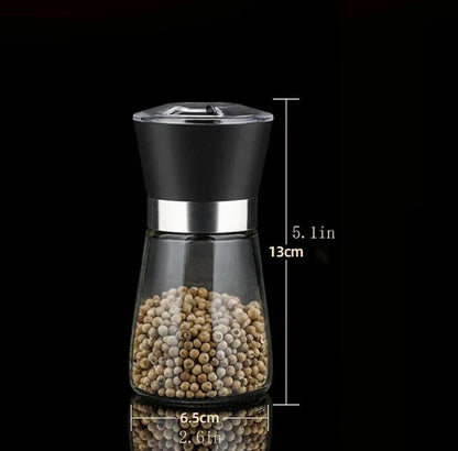 Elegant stainless steel pepper grinder with a clear glass body, shown set to a coarse grind, being used to season a dish of fresh food on a wooden counter.