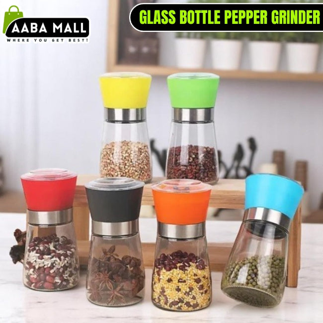 Elegant stainless steel pepper grinder with a clear glass body, shown set to a coarse grind, being used to season a dish of fresh food on a wooden counter.