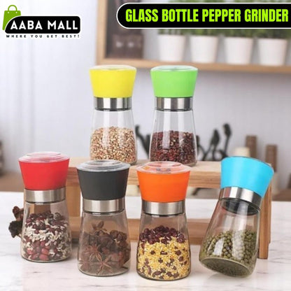 Elegant stainless steel pepper grinder with a clear glass body, shown set to a coarse grind, being used to season a dish of fresh food on a wooden counter.
