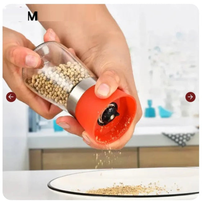 Elegant stainless steel pepper grinder with a clear glass body, shown set to a coarse grind, being used to season a dish of fresh food on a wooden counter.