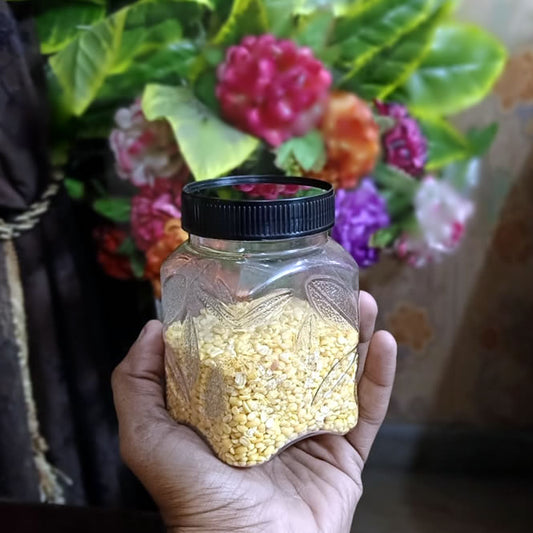 Hand holding a leaf jar with black lid filled with yellow seeds, with a blurred floral background.