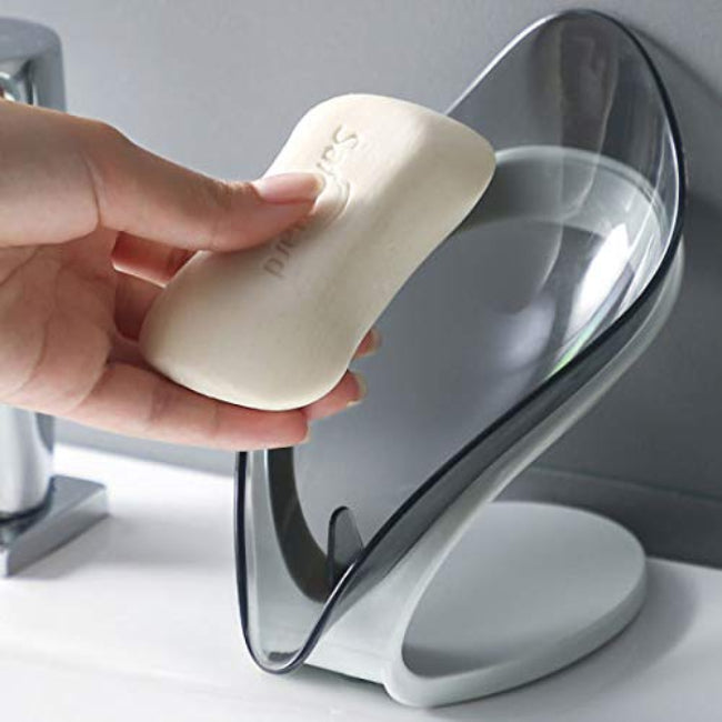 A green leaf-shaped self-draining soap dish holder with a suction cup base, shown on a sink edge, channeling water away from the soap bar.