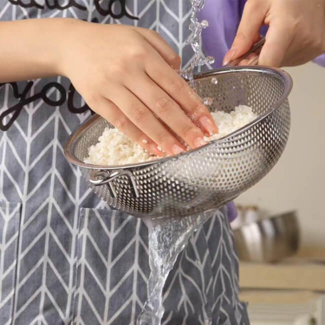 Person washing clothes in a metal strainer with water in a kitchen setting.