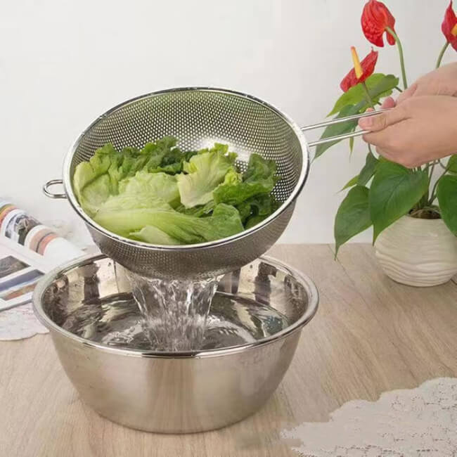 Person washing lettuce in a metal strainer over a bowl on a wooden table.