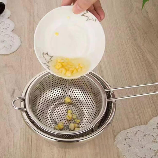 Person pouring contents from a white bowl into a metal strainer on a wooden surface.