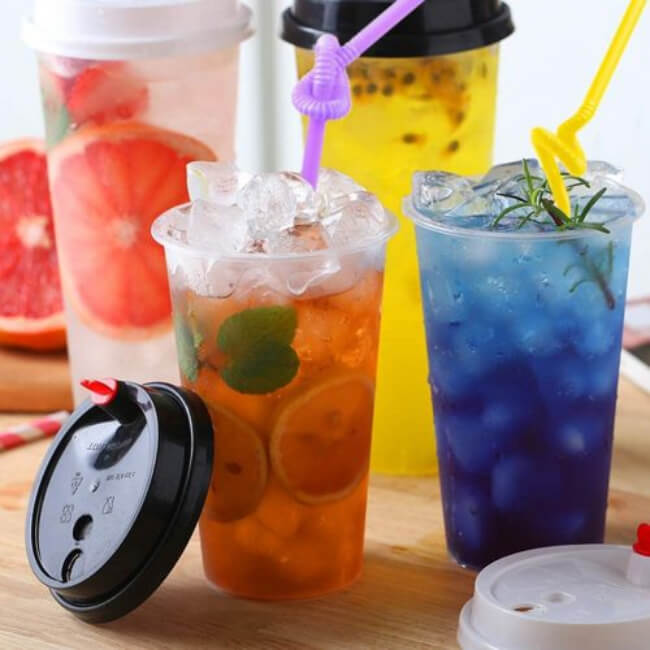 Four colorful drinks in clear plastic cups with lids and straws on a wooden surface.