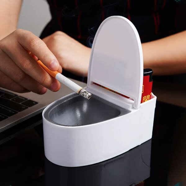 Toilet-shaped ashtray with a hand lighting a cigarette, on a gray background.